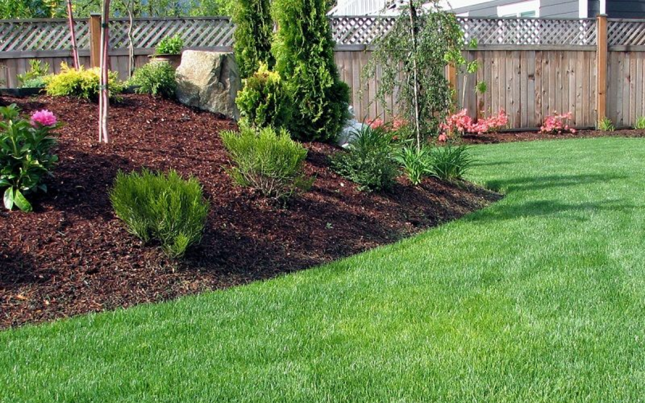 Gepflegter Garten mit grüner Rasenfläche, gemulchtem Blumenbeet, Sträuchern und einem Holzzaun