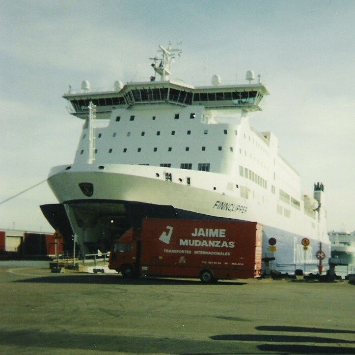 Un gran ferry blanco de pasajeros atracado junto a un contenedor rojo de Jaime Mudanzas en un puerto.