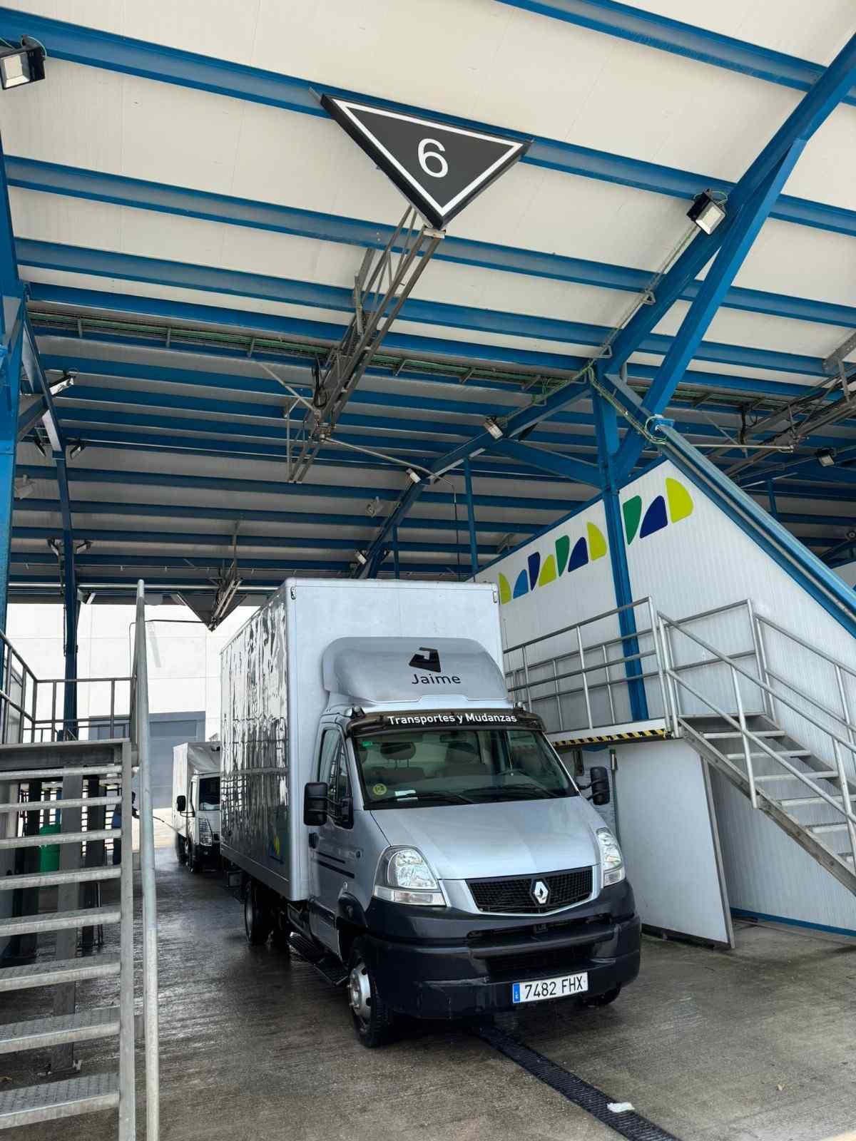 Un camión de reparto blanco estacionado bajo un muelle de carga con techo azul, junto a unas escaleras y barandillas.