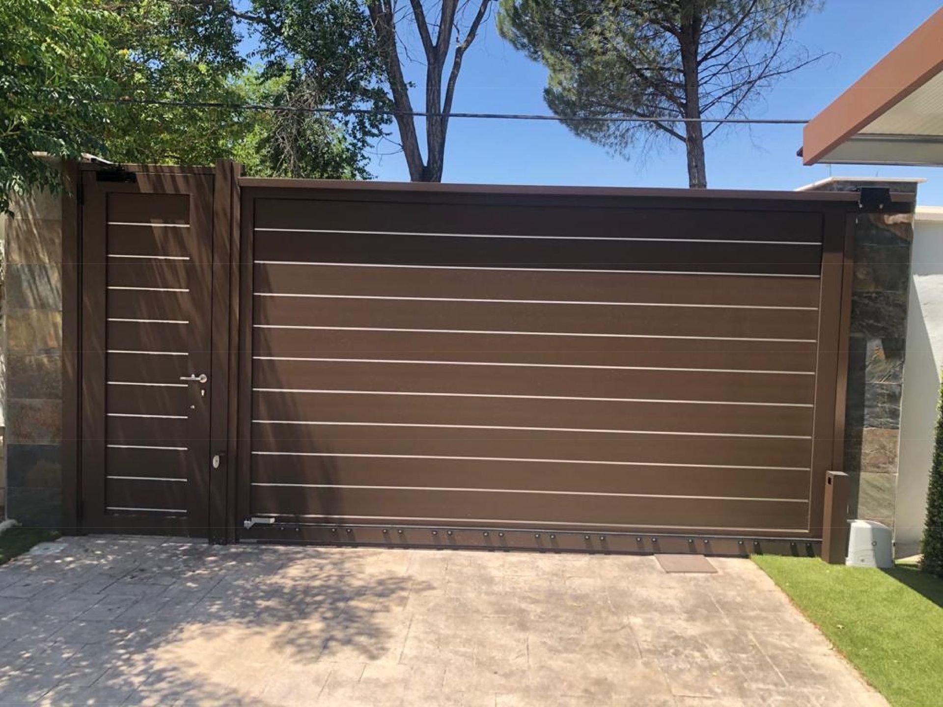 Una puerta corrediza marrón se abre a un camino de entrada con árboles al fondo.