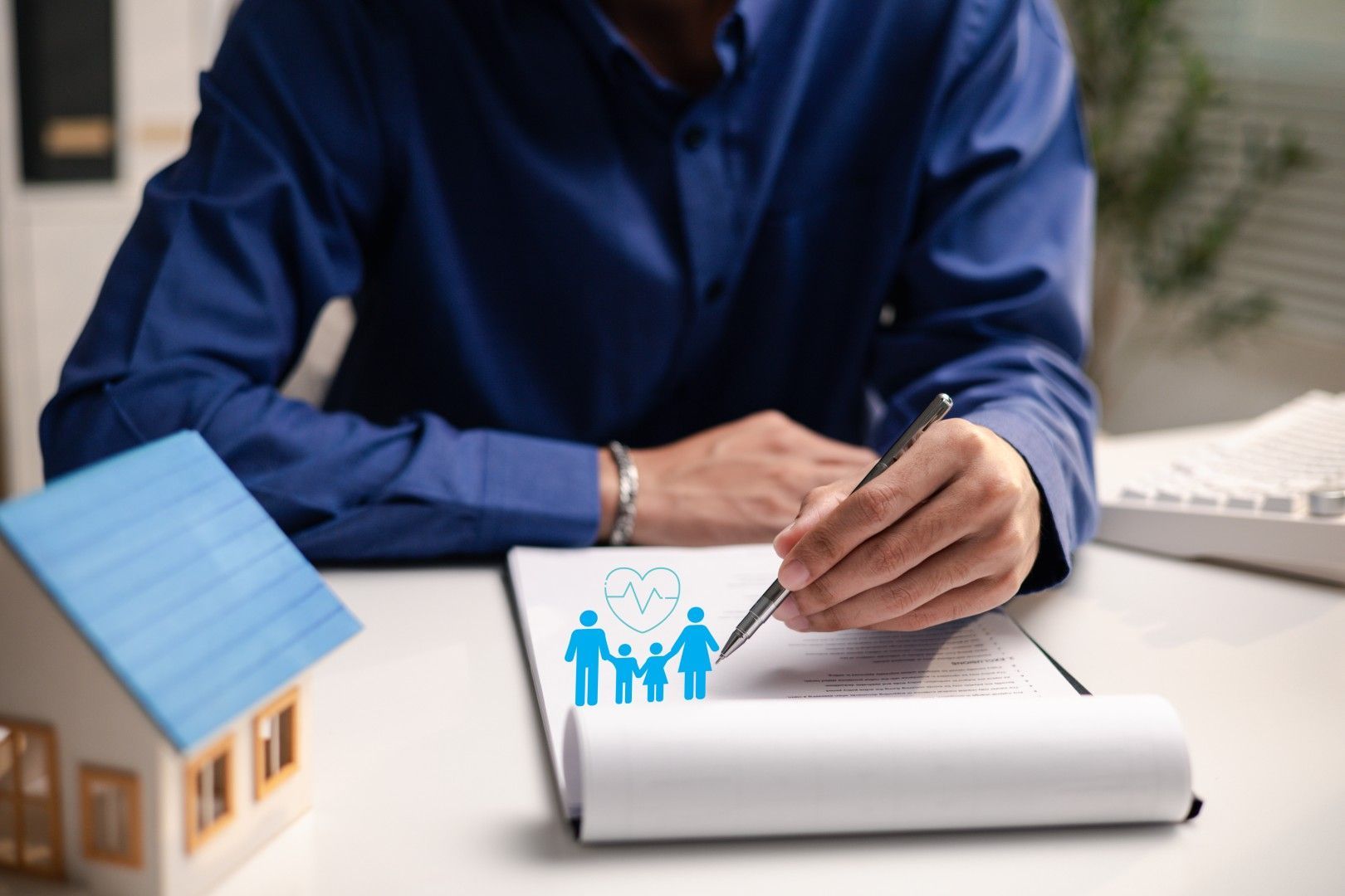 Personne en chemise bleue signant un document à côté d’une maquette de maison et d’une icône représentant une famille, sur un bureau