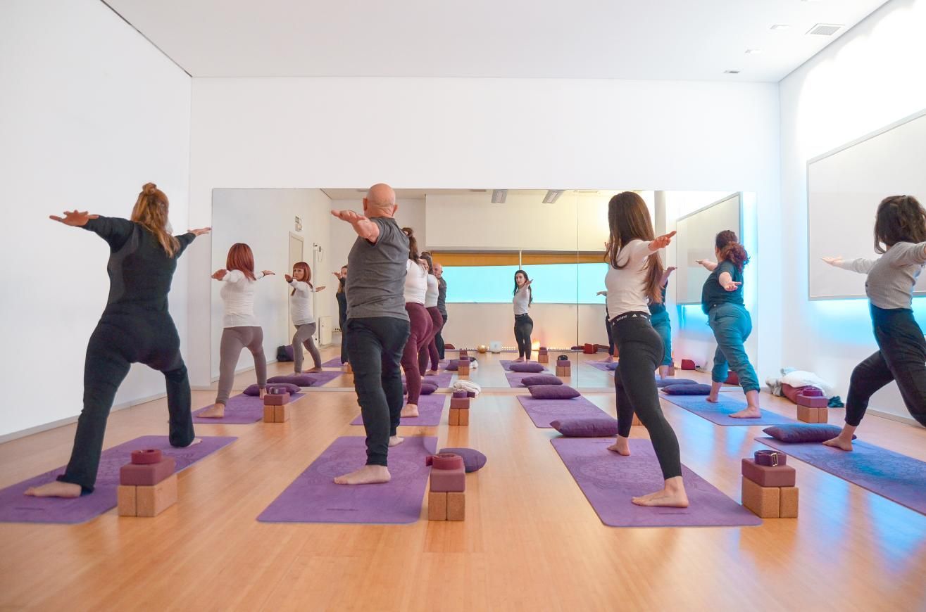 Dos mujeres en poses de yoga en un estudio con un espejo, con los brazos levantados y sentadas sobre colchonetas.