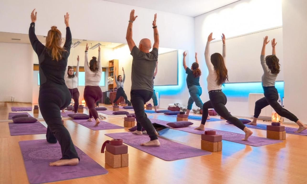 Mujeres en clase de yoga, sentadas, con los brazos en alto. Colchonetas moradas sobre suelo de madera.