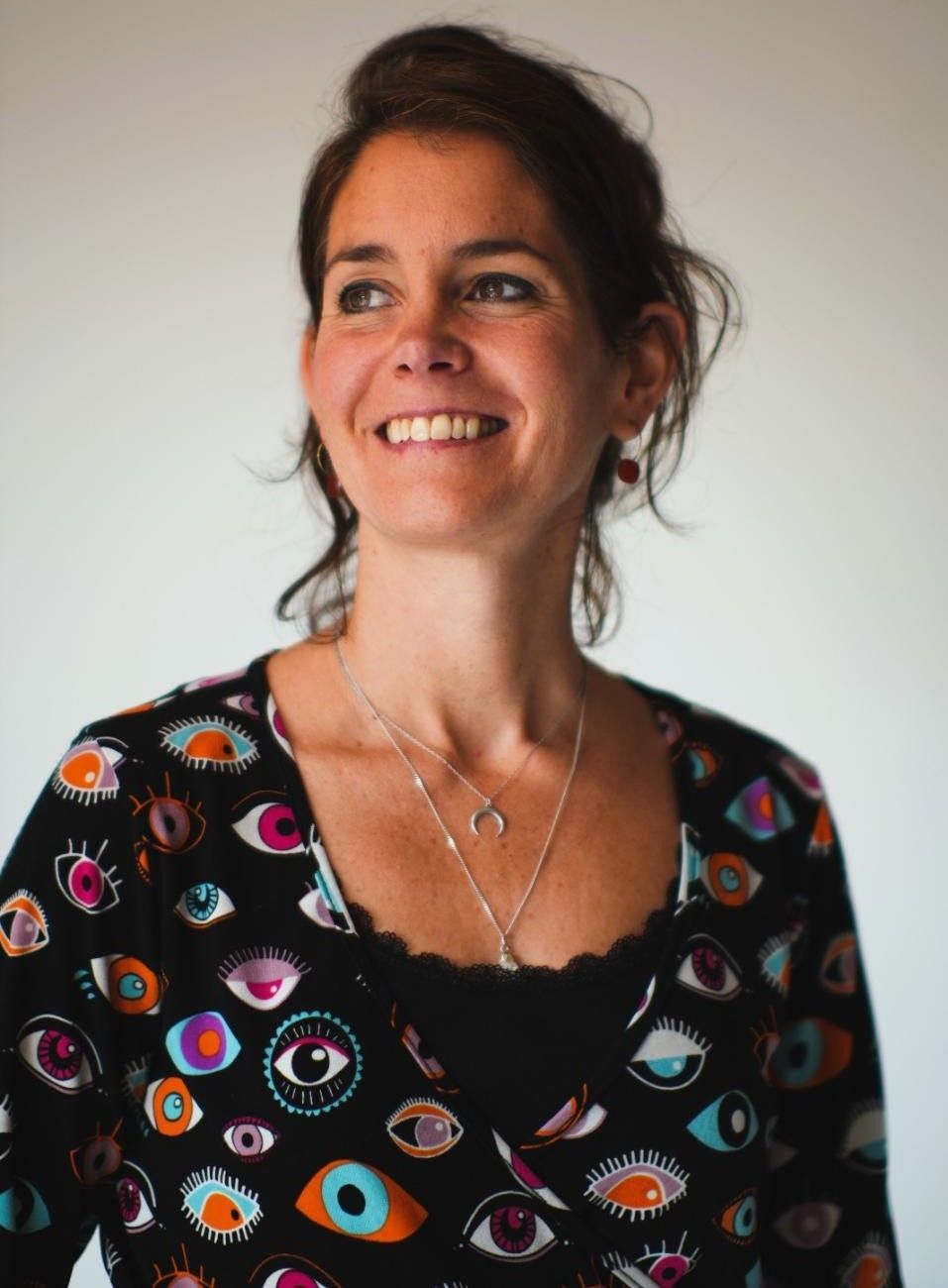 Una mujer sonríe mientras mira hacia arriba, viste una camisa negra con dibujos de ojos y un collar de plata.