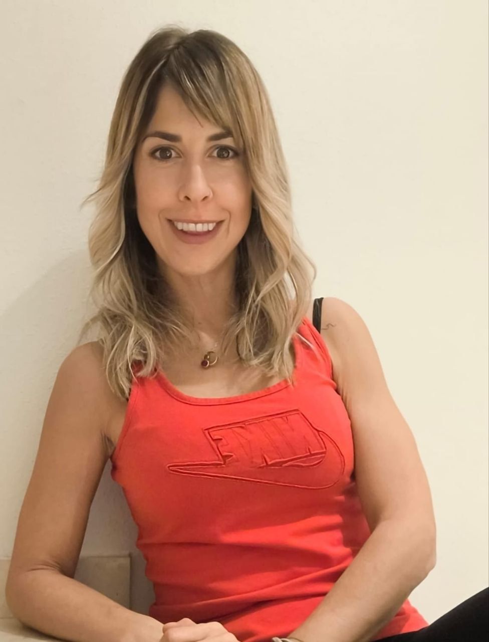 Una mujer con una camiseta roja sonríe, apoyada contra una pared blanca.