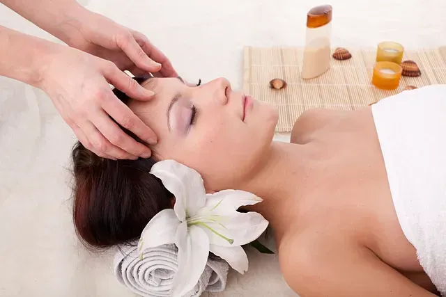 Mujer recibiendo un masaje de cabeza en un spa, con los ojos cerrados, una toalla blanca, un lirio y velas cerca.