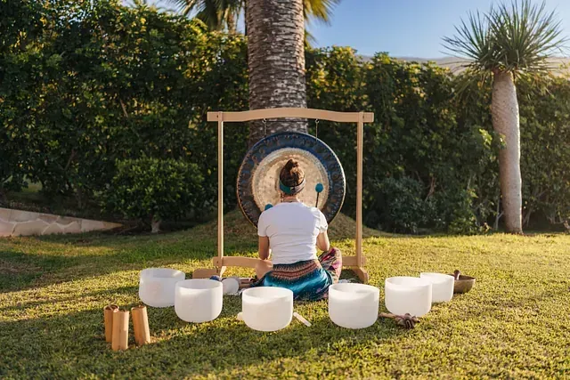 Persona de espaldas, tocando un gran gong en un jardín iluminado por el sol, con cuencos cantores de cristal dispuestos delante.