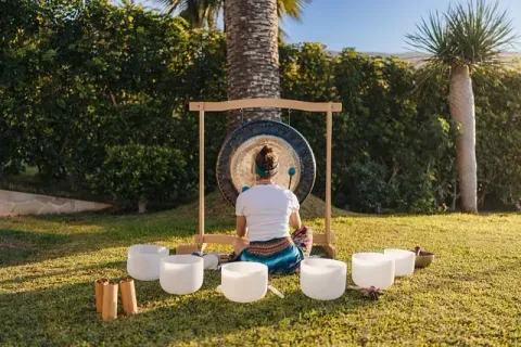 Persona de espaldas, tocando un gran gong en un jardín iluminado por el sol, con cuencos cantores de cristal dispuestos delante.