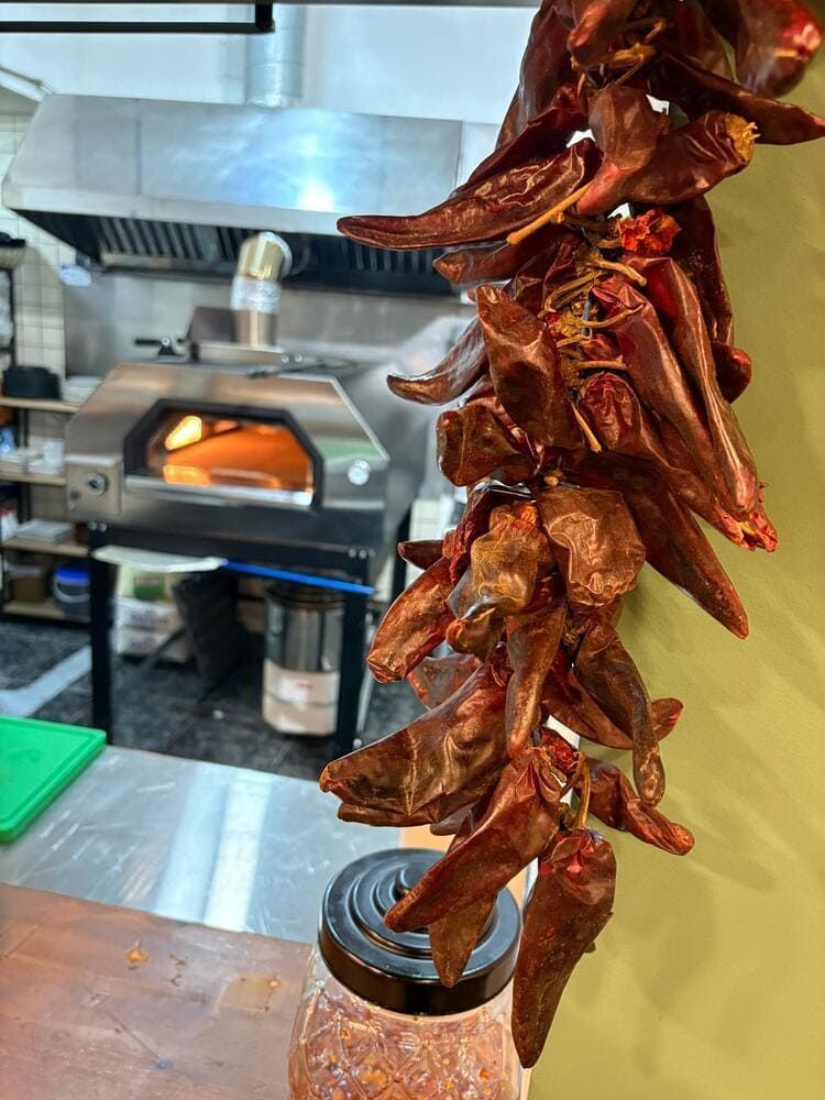 Chiles rojos secos colgando en una cocina industrial, con un horno de pizza al fondo.