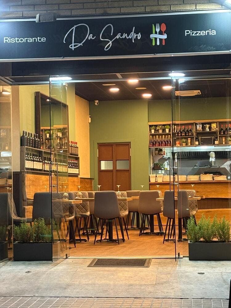 Fachada de pizzería italiana moderna con entrada de cristal, iluminación cálida y mesas vacías en el interior.