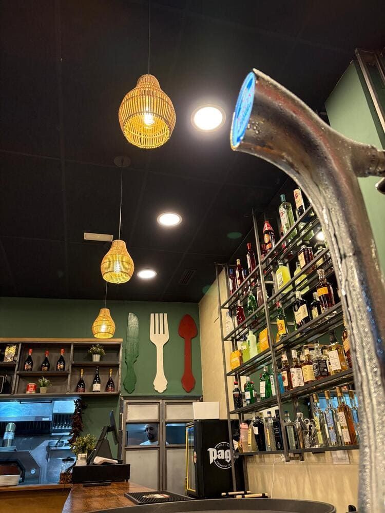 Interior acogedor de una cafetería con luces colgantes, estantes con botellas y un gran grifo metálico en primer plano.