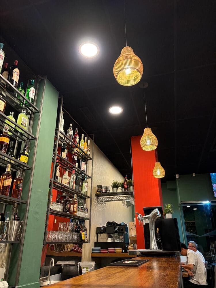 Interior de bar acogedor con estantes llenos de botellas, lámparas colgantes y una persona sentada en la barra.