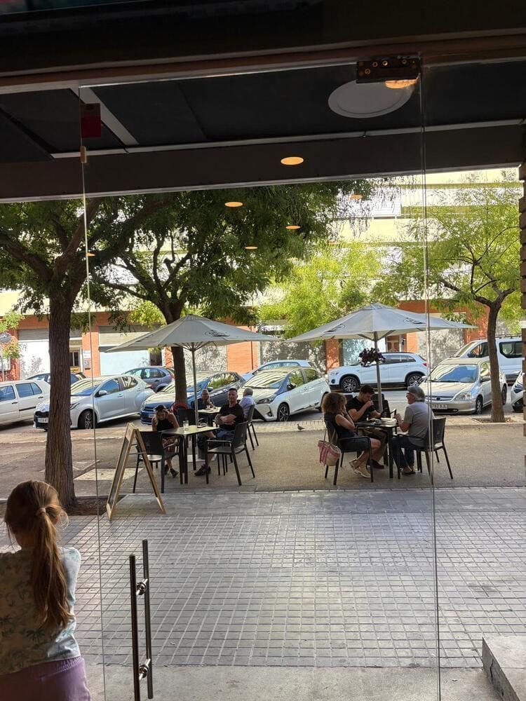 Terraza de una cafetería bajo un patio cubierto, con gente sentada en las mesas y coches aparcados más allá de la calle.