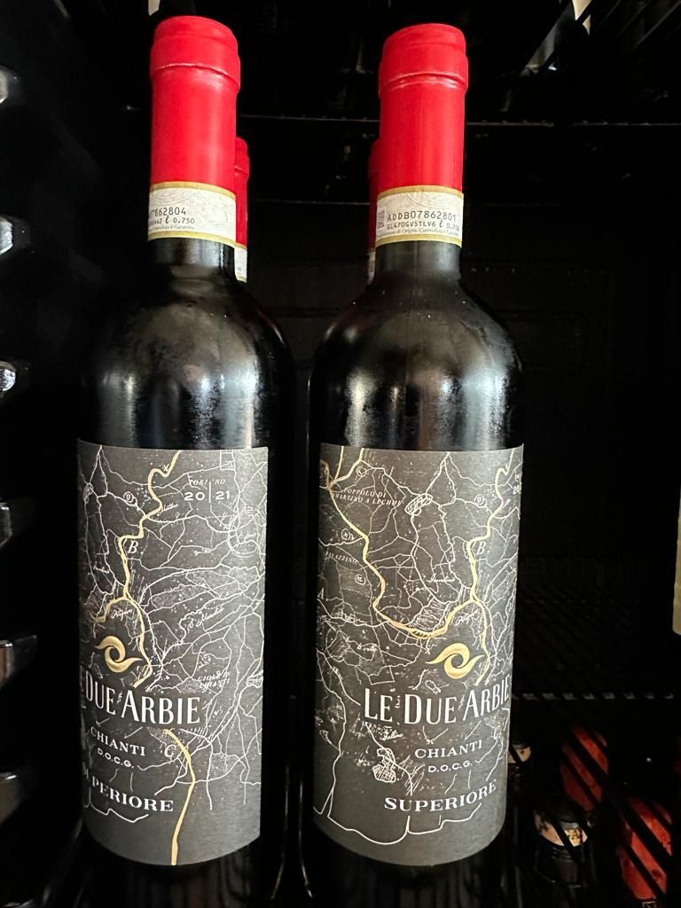 Dos botellas de vino tinto oscuro con tapones rojos y etiquetas negras sobre una superficie oscura.