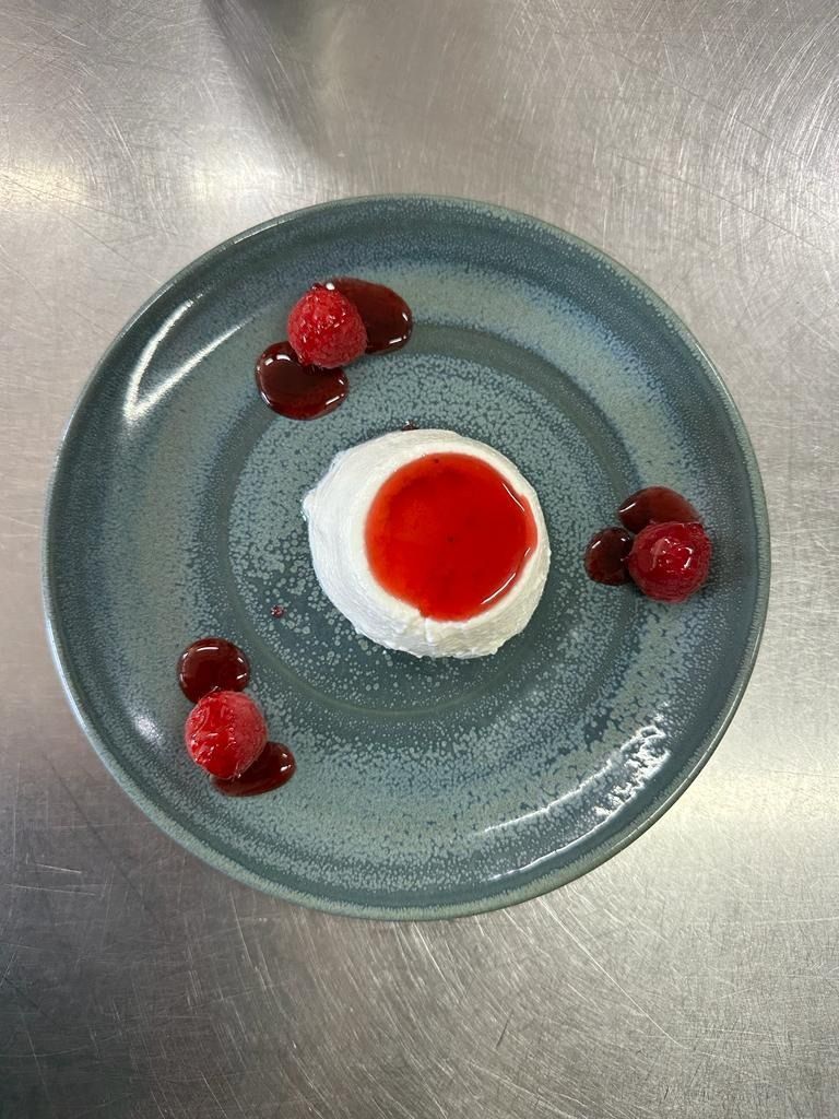 Plato azul con un postre blanco y rojo, adornado con frambuesas sobre una mesa de metal.