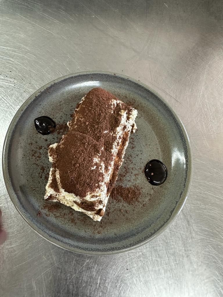 Una porción de postre en un plato transparente con cacao en polvo y dos gotas de salsa de chocolate sobre una mesa de metal.