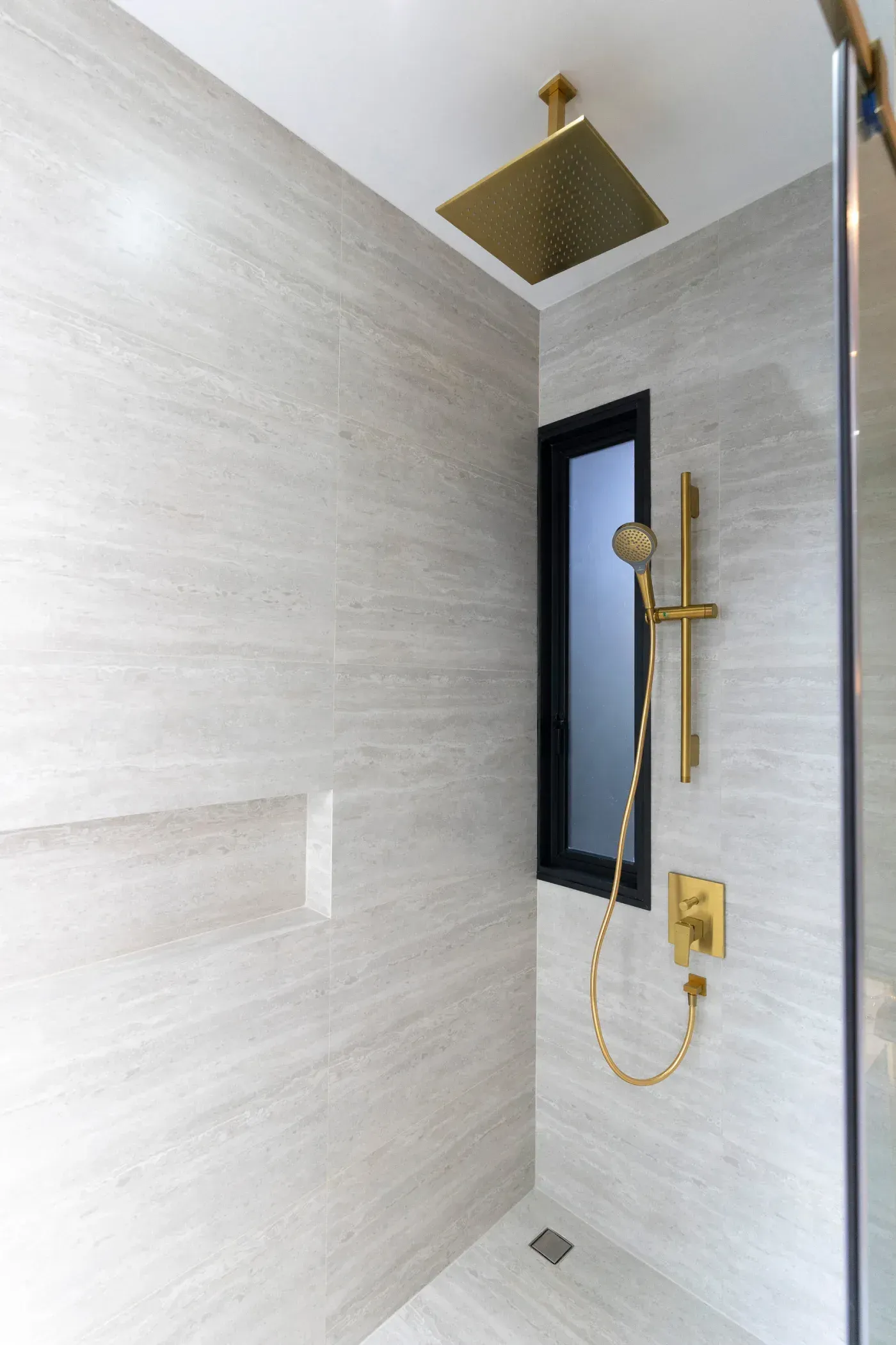 Ducha moderna con accesorios dorados y paredes de azulejos gris claro.