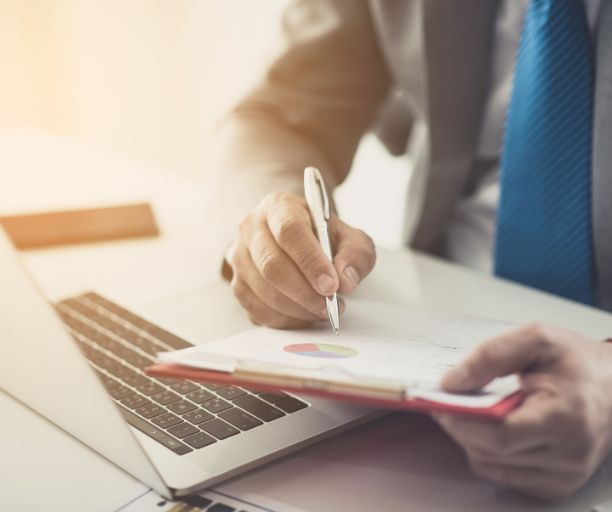 A man in a suit and tie is writing on a clipboard in front of a laptop computer.