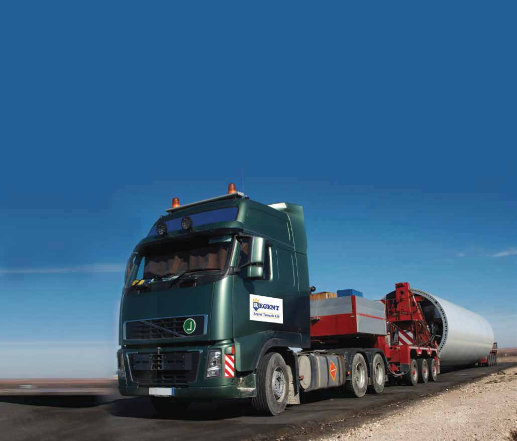 A green semi truck is driving down a road carrying a large wind turbine.