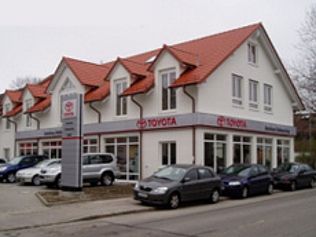 Toyota-Händlergebäude mit draußen geparkten Autos. Rotes Dach, weiße Fassade, bewölkter Tag.