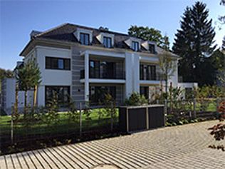 Modernes, weißes, mehrstöckiges Gebäude mit Balkonen, Dachgauben und einer Auffahrt aus grauem Stein; vor blauem Himmel und Bäumen.