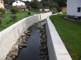 Ein betonierter Bach fließt durch ein Wohngebiet, im Hintergrund ein grasbewachsenes Ufer und Häuser.