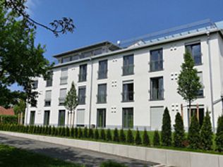 Weißes Mehrfamilienhaus mit vielen Fenstern und Balkonen; davor eine Reihe kleiner Bäume.