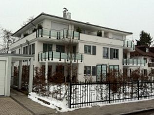 Weißes, modernes Apartmentgebäude mit Glasbalkonen, Schnee und schwarzem Zaun.