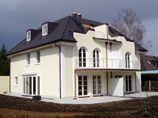 Zweistöckiges beiges Haus mit dunklem Dach und Balkon vor einem bewölkten Himmel.