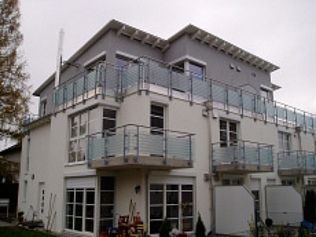 Dreistöckiges weißes Gebäude mit grauem Dach und Balkonen mit Glasgeländern. Bewölkter Tag.