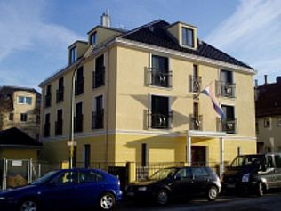 Gelbes Gebäude mit Balkonen, davor geparkten Autos und einer Flagge.