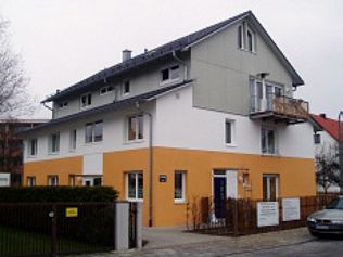 Dreistöckiges Gebäude mit orangefarbenen und weißen Wänden, grauem Dach, Balkon und einem davor geparkten Auto.