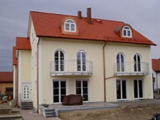 Cremefarbenes Haus mit rotem Dach, Bogenfenstern, Balkonen und einer kleinen Veranda mit Stufen. Bewölkter Tag.