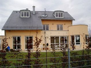 Gelbes zweistöckiges Haus mit grauem Dach, großen Fenstern und einem geschwungenen Seitenteil, hinter einem Zaun.