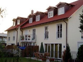 Beiges zweistöckiges Gebäude mit rotem Ziegeldach, Dachgauben und Sitzgelegenheiten auf der Terrasse auf einem grünen Rasen.