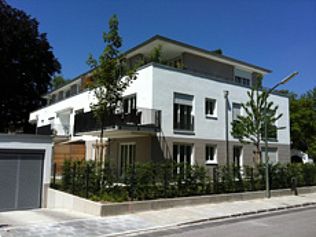 Modernes, weiß-graues Apartmentgebäude mit Dachterrasse, Bäumen und klarem, blauem Himmel.