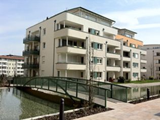 Modernes Apartmentgebäude mit Balkonen über einem Wasserspiel, verbunden durch eine Fußgängerbrücke.