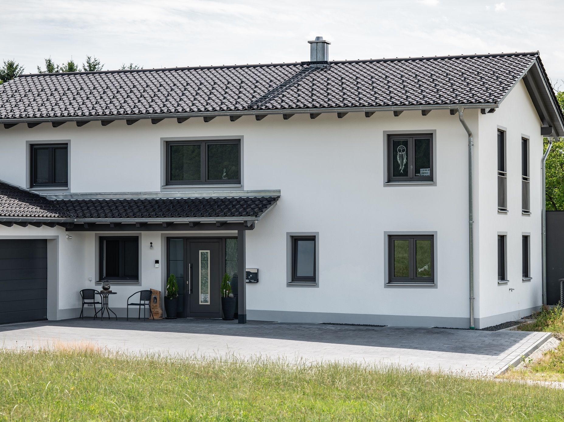 Weißes zweistöckiges Haus mit dunkelgrauen Zierleisten und Dach, grau gepflasterte Auffahrt.