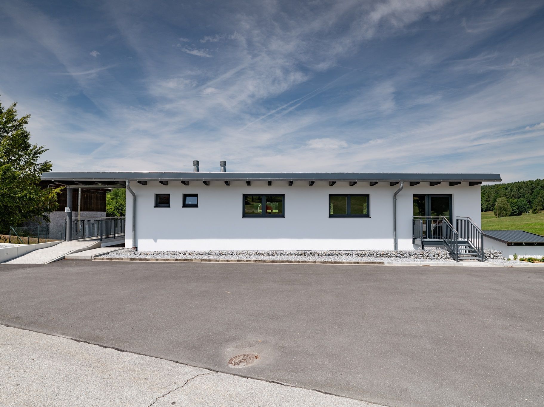 Weißes Gebäude mit schwarz gerahmten Fenstern und Türen; graues Dach. Bewölkter Himmel, asphaltierte Auffahrt, grüne Landschaft.