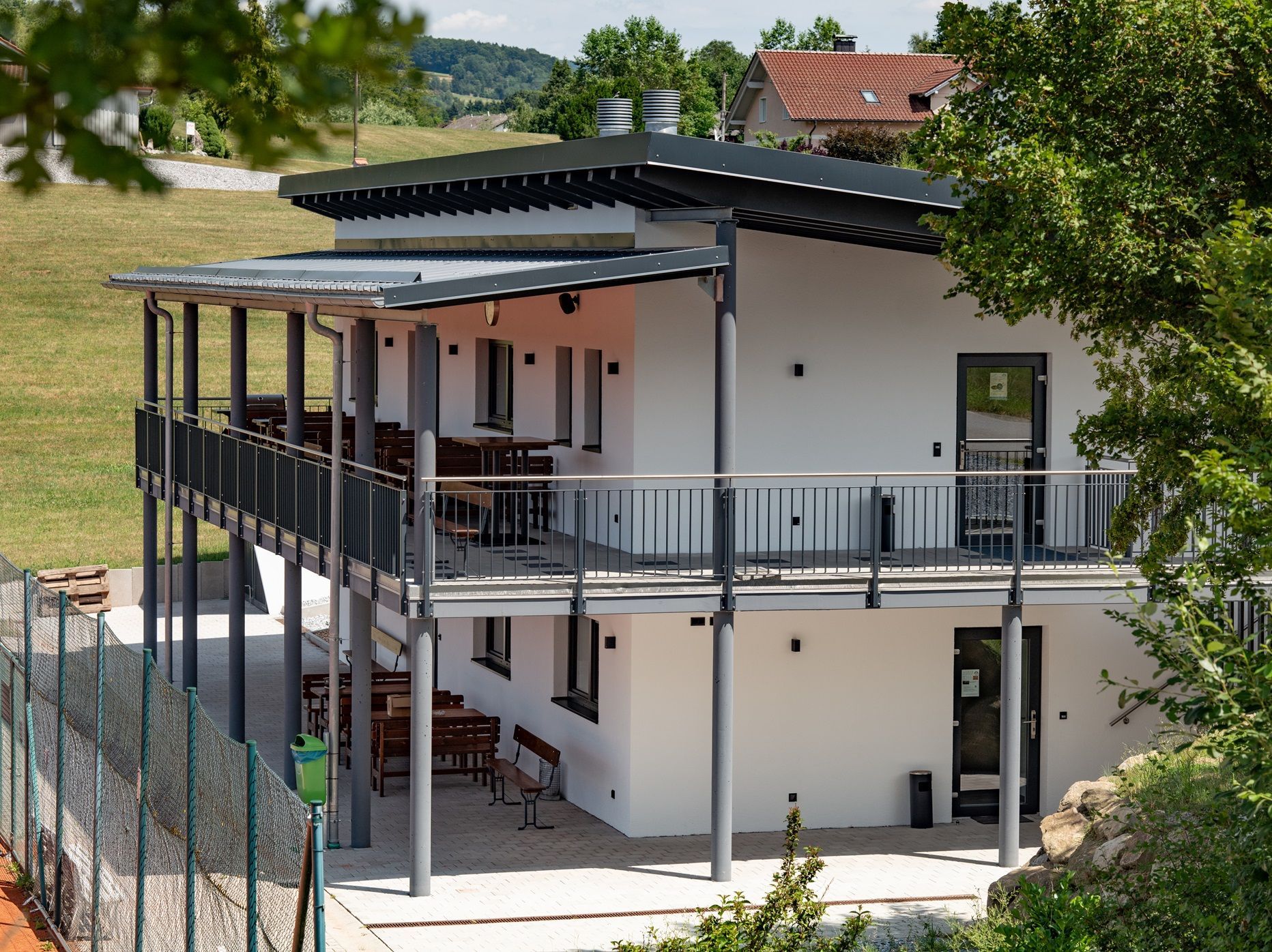 Zweistöckiges weißes Gebäude mit Balkon und Blick auf einen Tennisplatz; grünes Laub im Vordergrund.