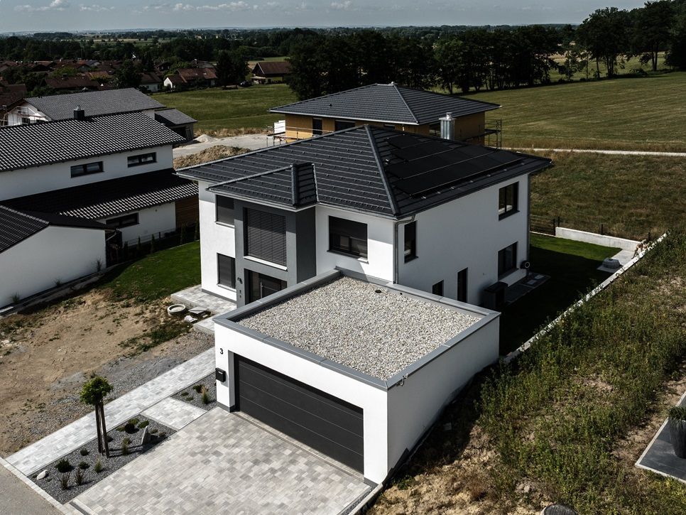 Modernes zweistöckiges Haus mit dunklem Dach, Garage und umgebendem Grün. Weiße Außenseite und graue Akzente.