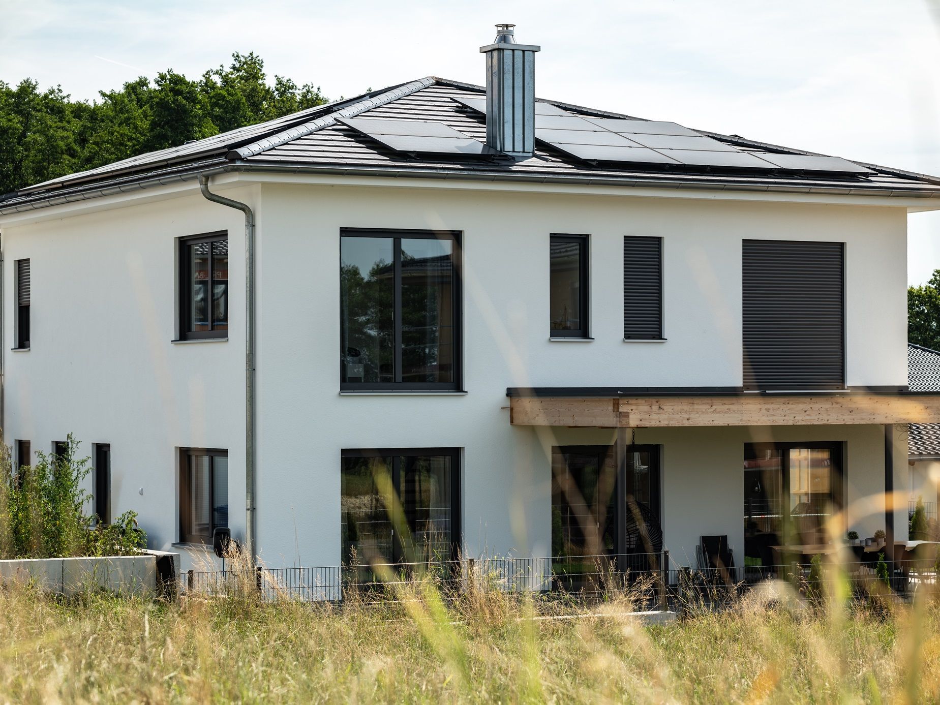 Weißes zweistöckiges Haus mit schwarz gerahmten Fenstern, Solarzellen auf dem Dach und einer überdachten Terrasse.