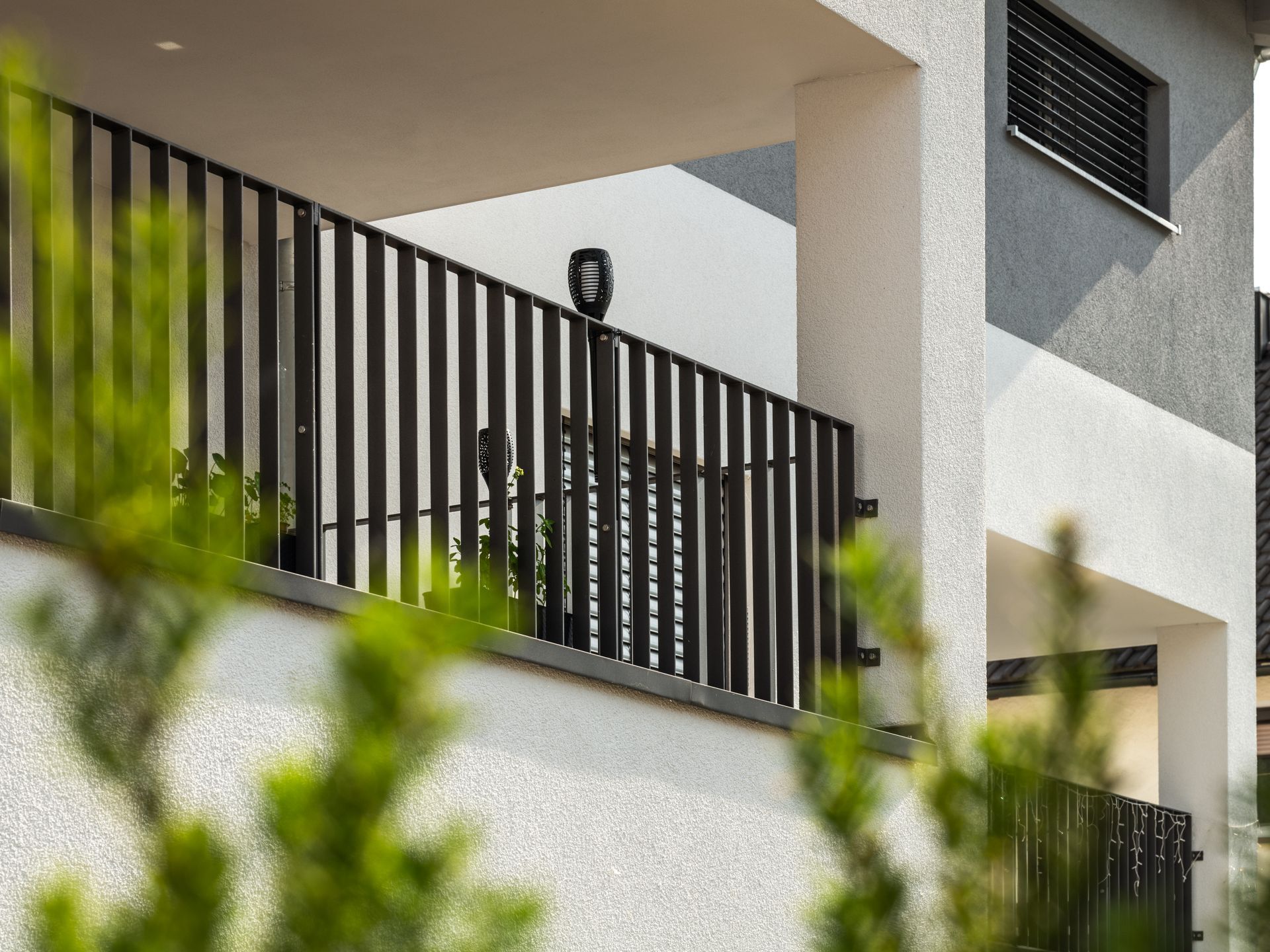 Moderne Gebäudefassade mit schwarzem Geländer und Balkon, teilweise verdeckt durch grünes Laub.