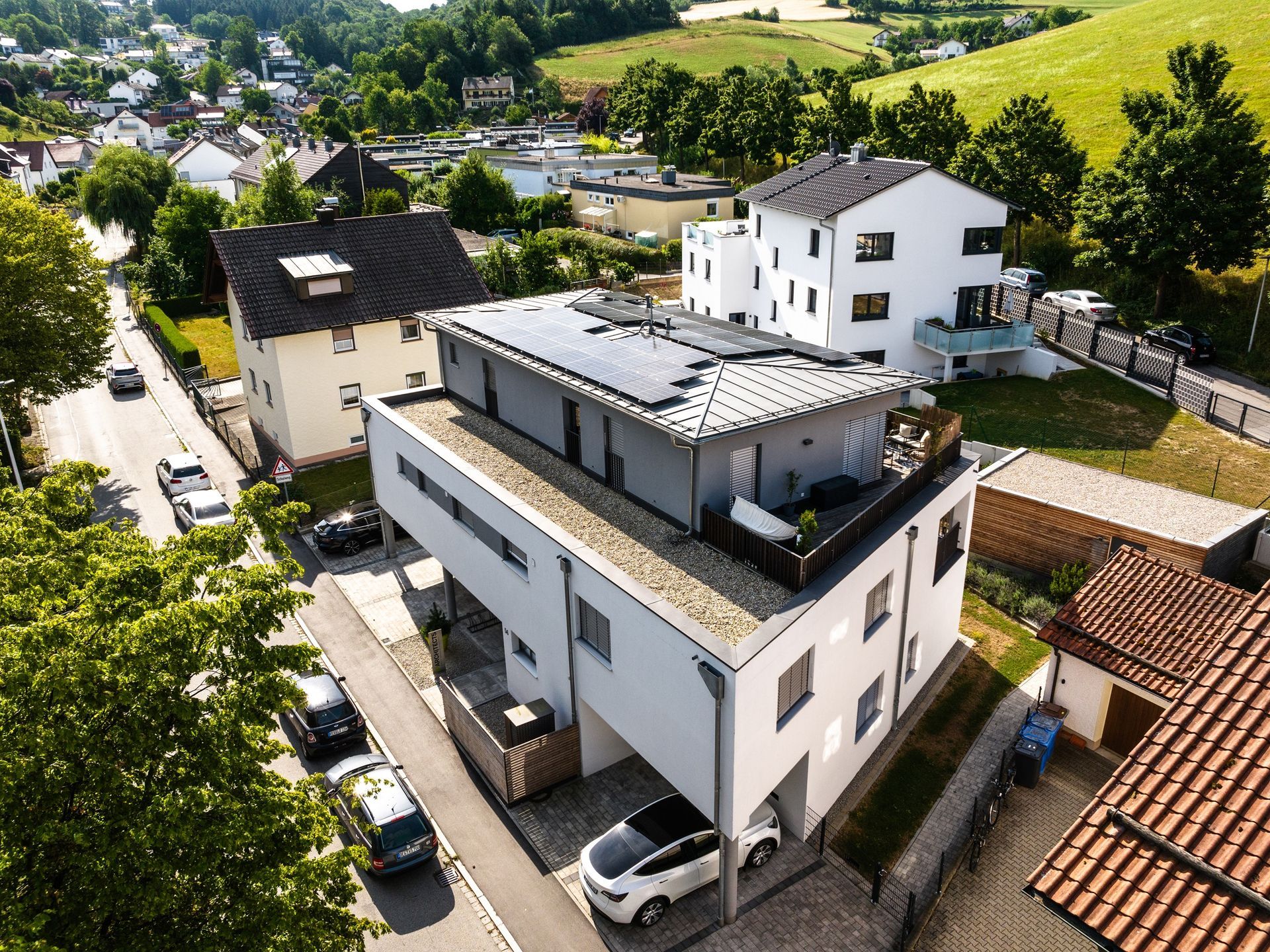 Modernes weißes mehrstöckiges Gebäude mit Solarzellen auf dem Dach; Häuser und grüne Landschaft im Hintergrund.