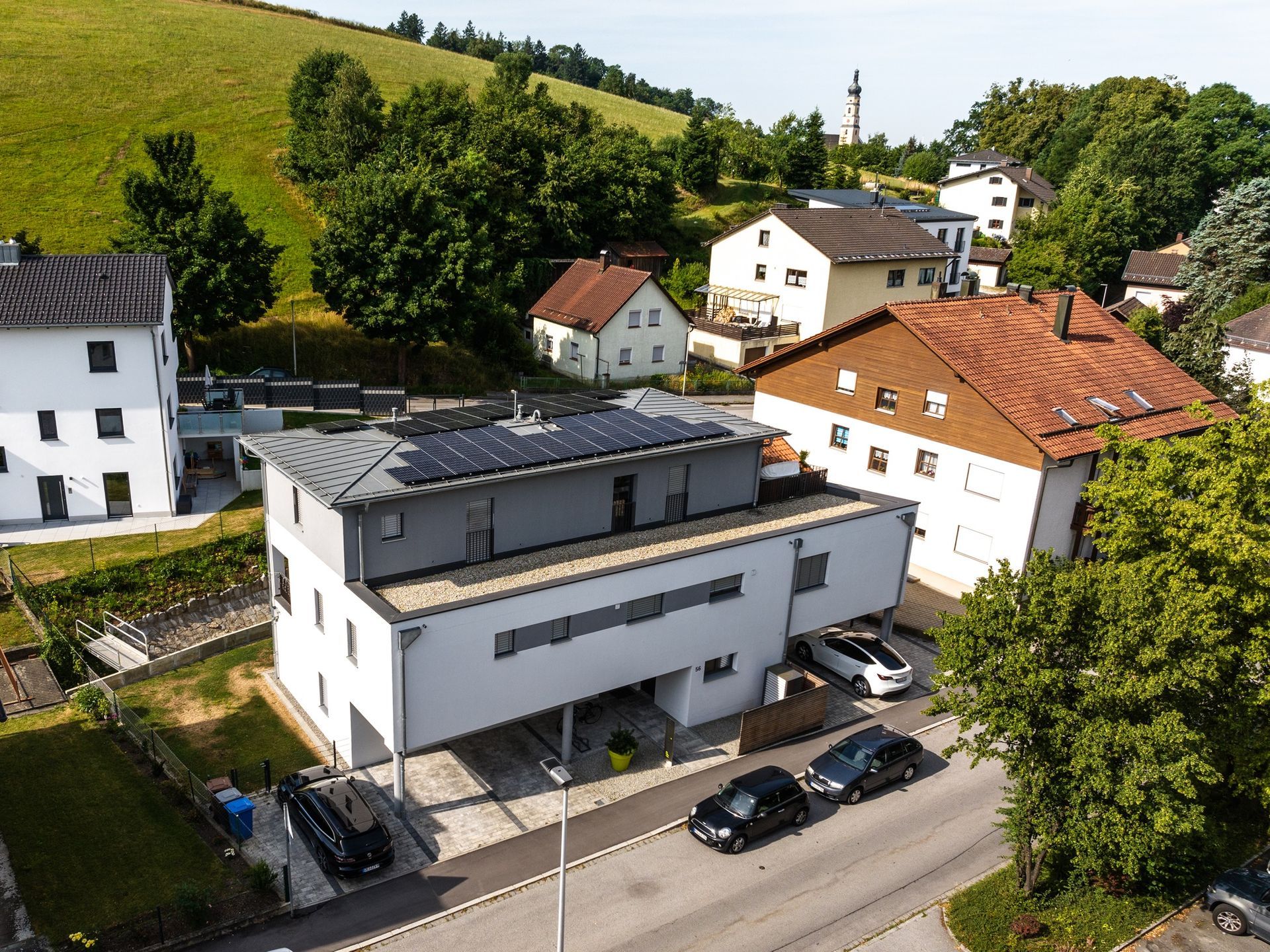 Modernes weißes Gebäude mit Solarmodulen und auf einer Straße geparkten Autos, Häusern und einem grünen Hügel im Hintergrund.