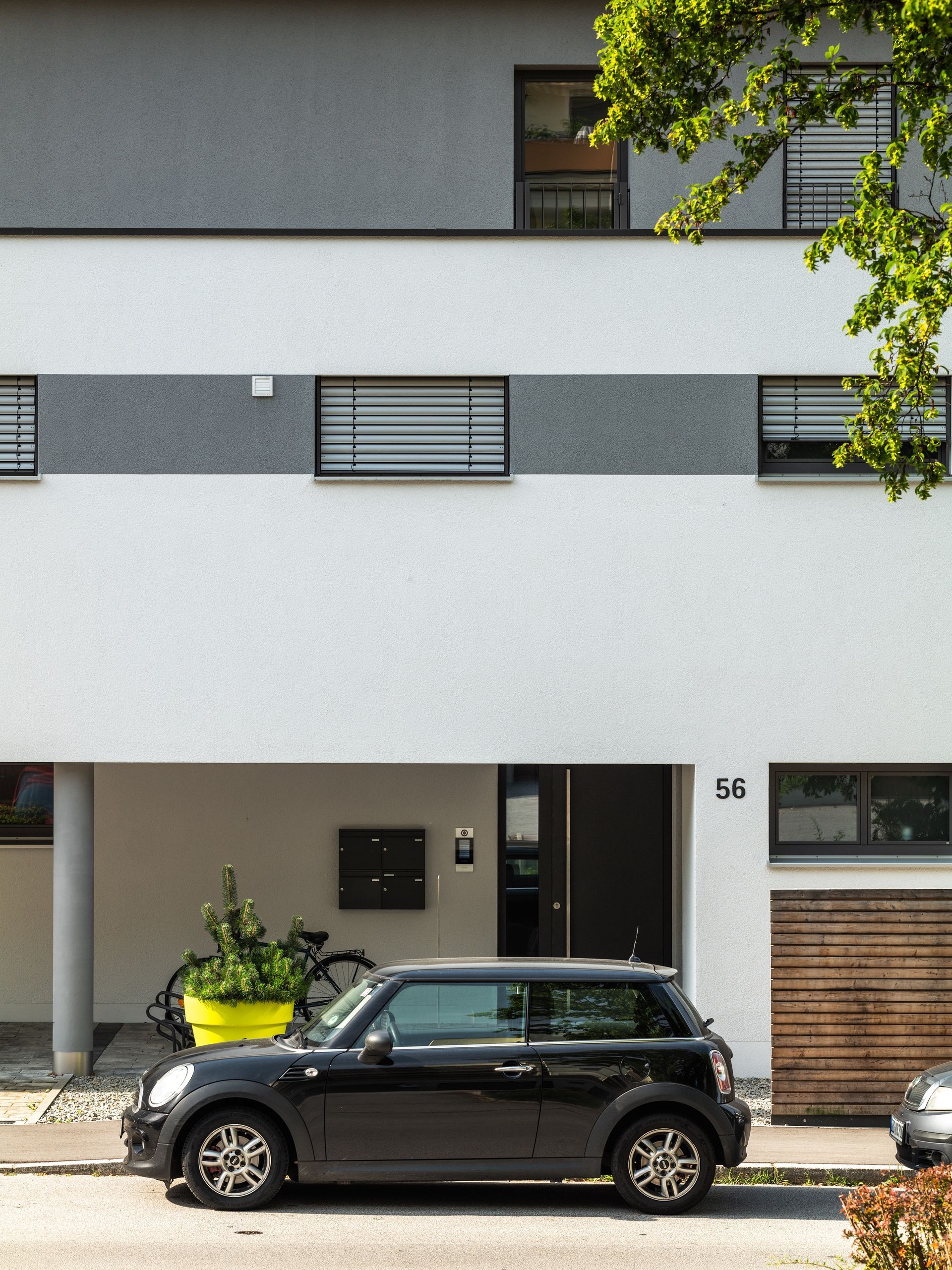 Schwarzer Mini Cooper parkt vor einem modernen weißen Gebäude mit horizontalen grauen Streifen und Ästen.