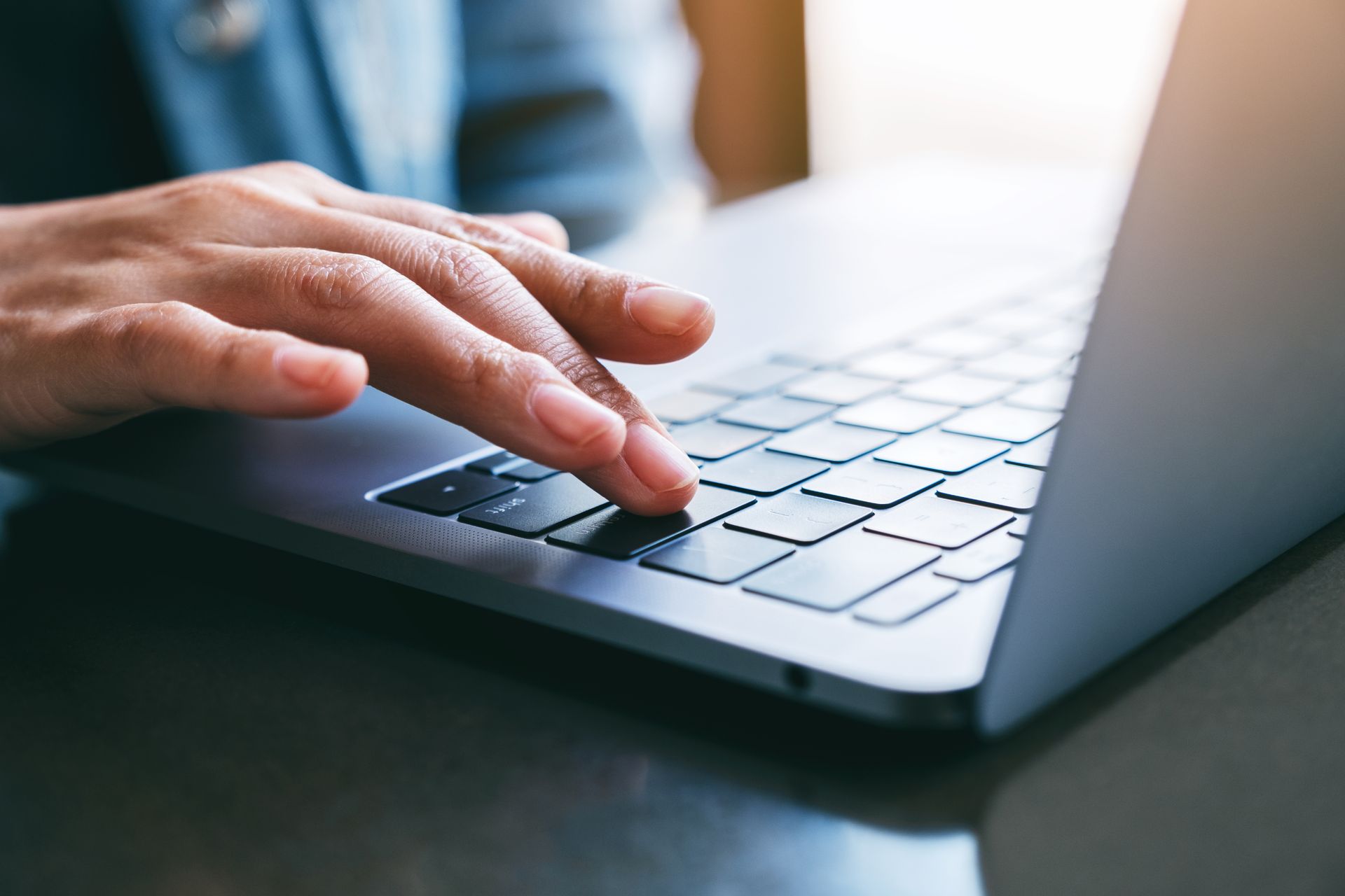 Eine Hand tippt auf einer silbernen Laptop-Tastatur neben einer blauen Tasse auf einem Tisch.