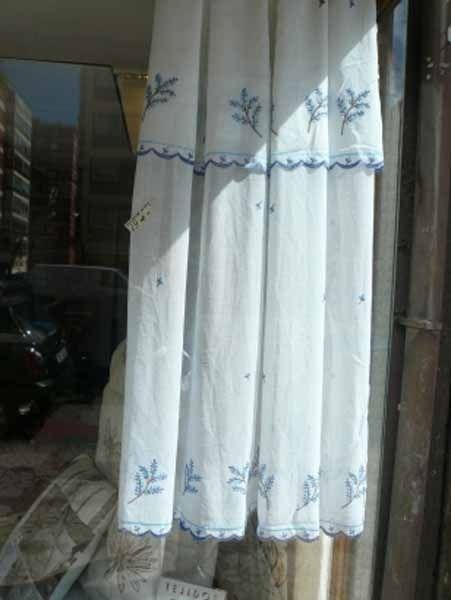 Un par de cortinas blancas con bordados azules cuelgan en una ventana.