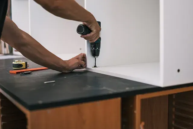 Un hombre está usando un taladro para atornillar un trozo de madera a un armario.