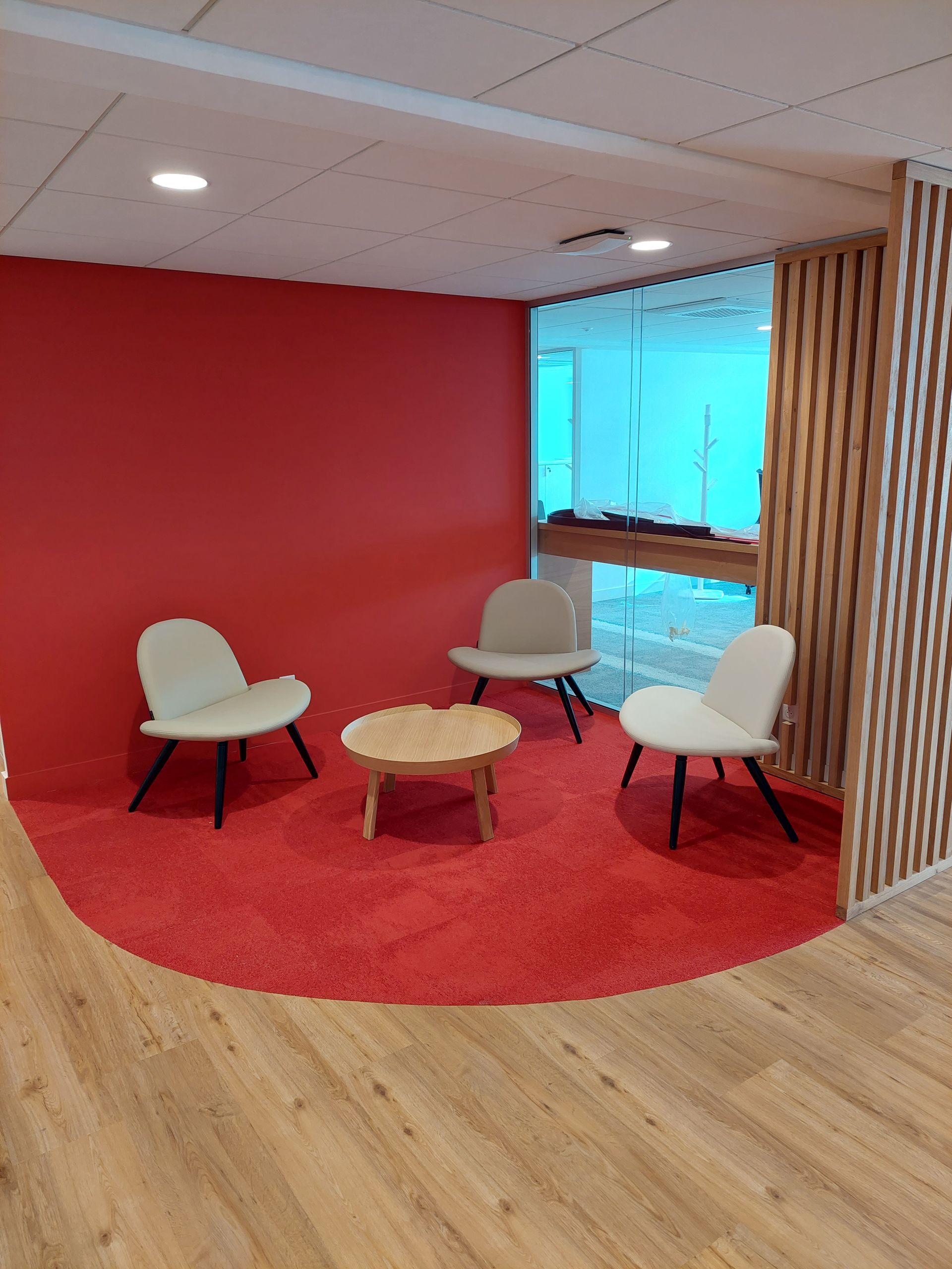 Espace d'attente confortable avec moquette rouge, trois chaises, petite table, mur rouge et éléments en bois.